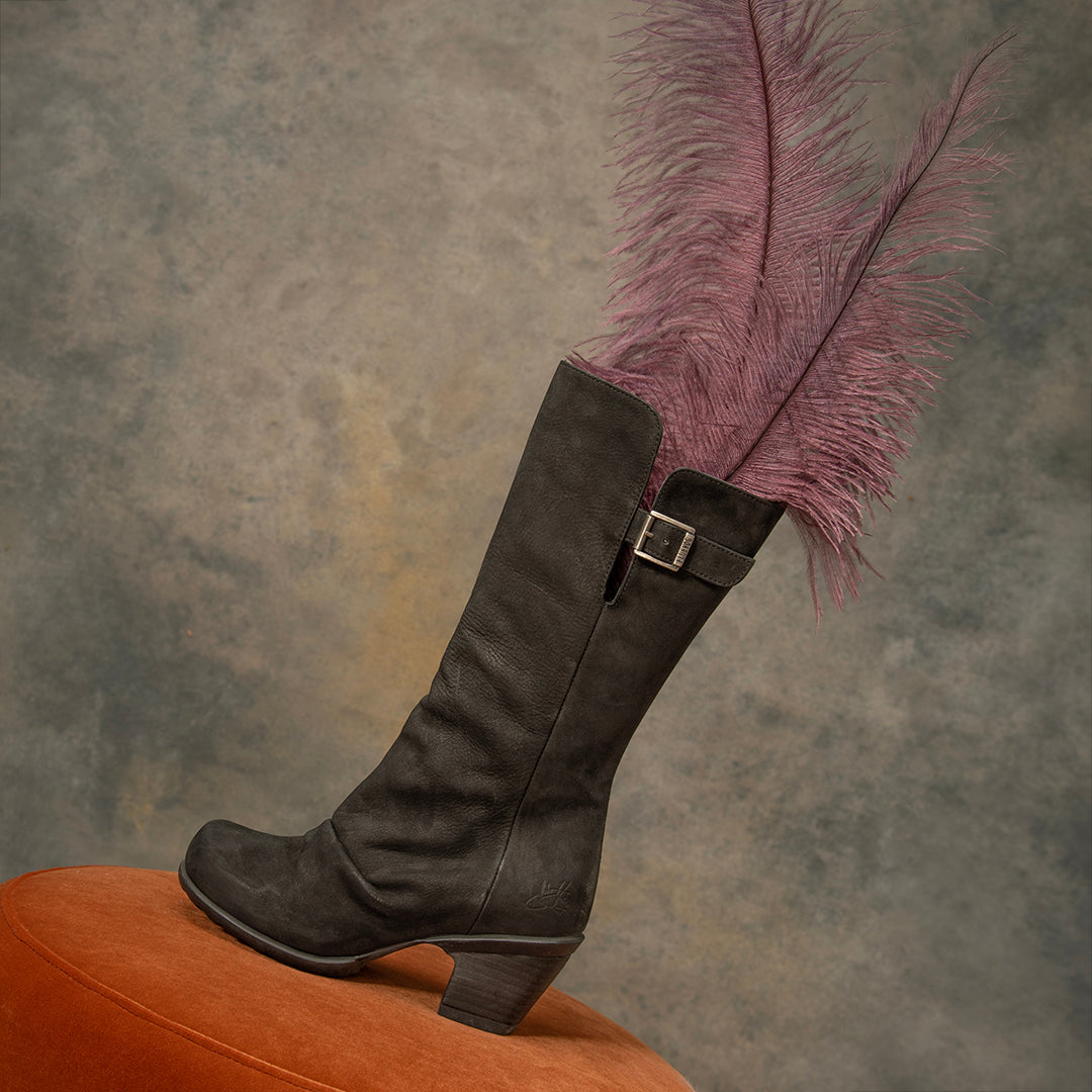 The Kanawa in black leather knee-high boot with a block heel and a decorative buckle strap near the top, styled with a long dusty purple feather and resting on an orange stool against a textured gray studio background.