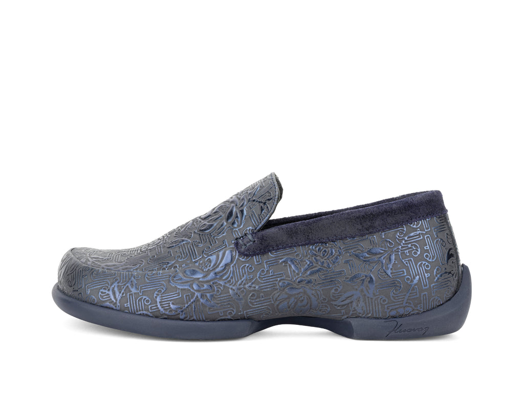 A slip-on loafer shoe with a textured, embossed pattern in slate blue and navy, featuring a dark navy plush collar and sole, shown from a side profile on a white background.