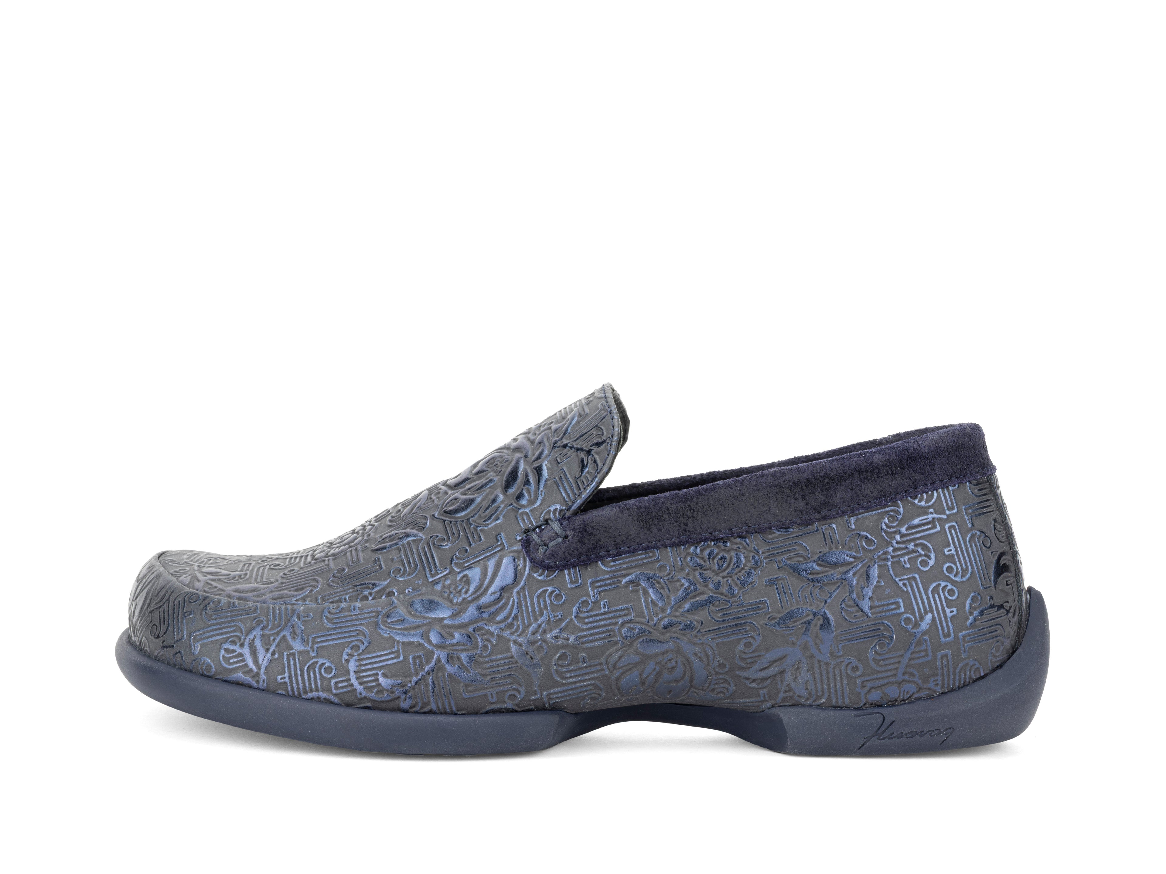 A slip-on loafer shoe with a textured, embossed pattern in slate blue and navy, featuring a dark navy plush collar and sole, shown from a side profile on a white background.